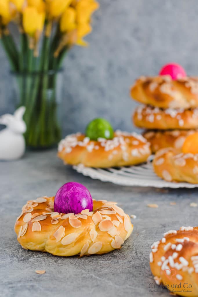 Osterkränze aus Dinkelmehl mit gefärbten Eiern und Narzissen im Hintergrund.