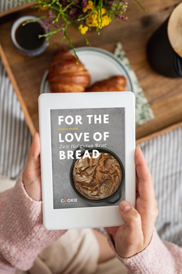 E-Book For the love of bread auf einem IPad in den Händen einer Frau mit Croissants im Hintergrund.