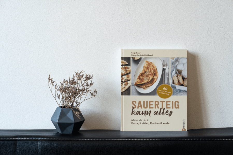 Backbuch und Kochbuch mit Sauerteig "Sauerteig kann alles" neben einer Vase mit Trockenblumen