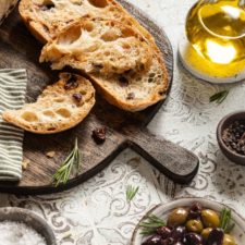 Mediterranes Olivenbrot aufgeschnitten auf einem Holzbrett mit einer Schale Oliven und einer Flasche Olivenöl.