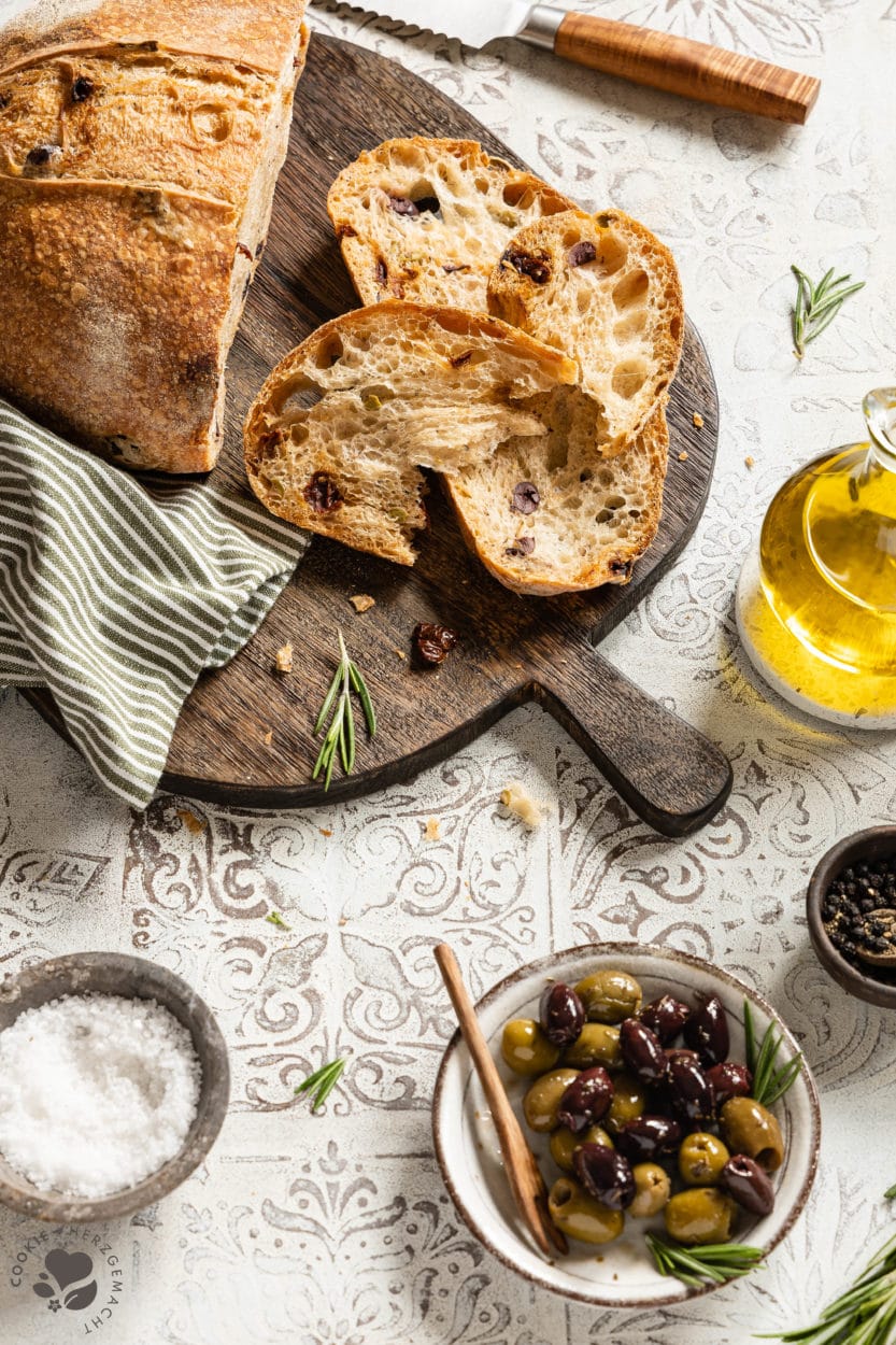Mediterranes Olivenbrot aufgeschnitten auf einem Holzbrett mit einer Schale Oliven und einer Flasche Olivenöl.