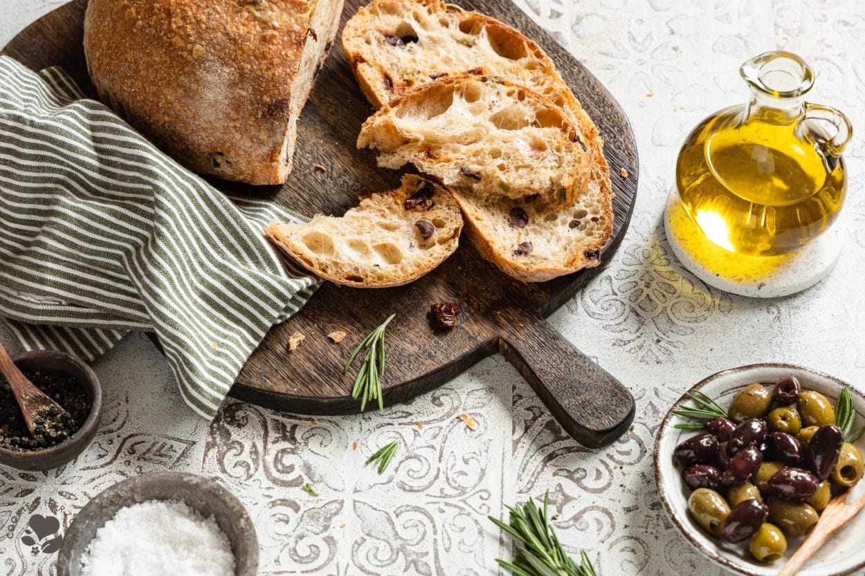 Mediterranes Olivenbrot aufgeschnitten auf einem Holzbrett mit einer Schale Oliven und einer Flasche Olivenöl.