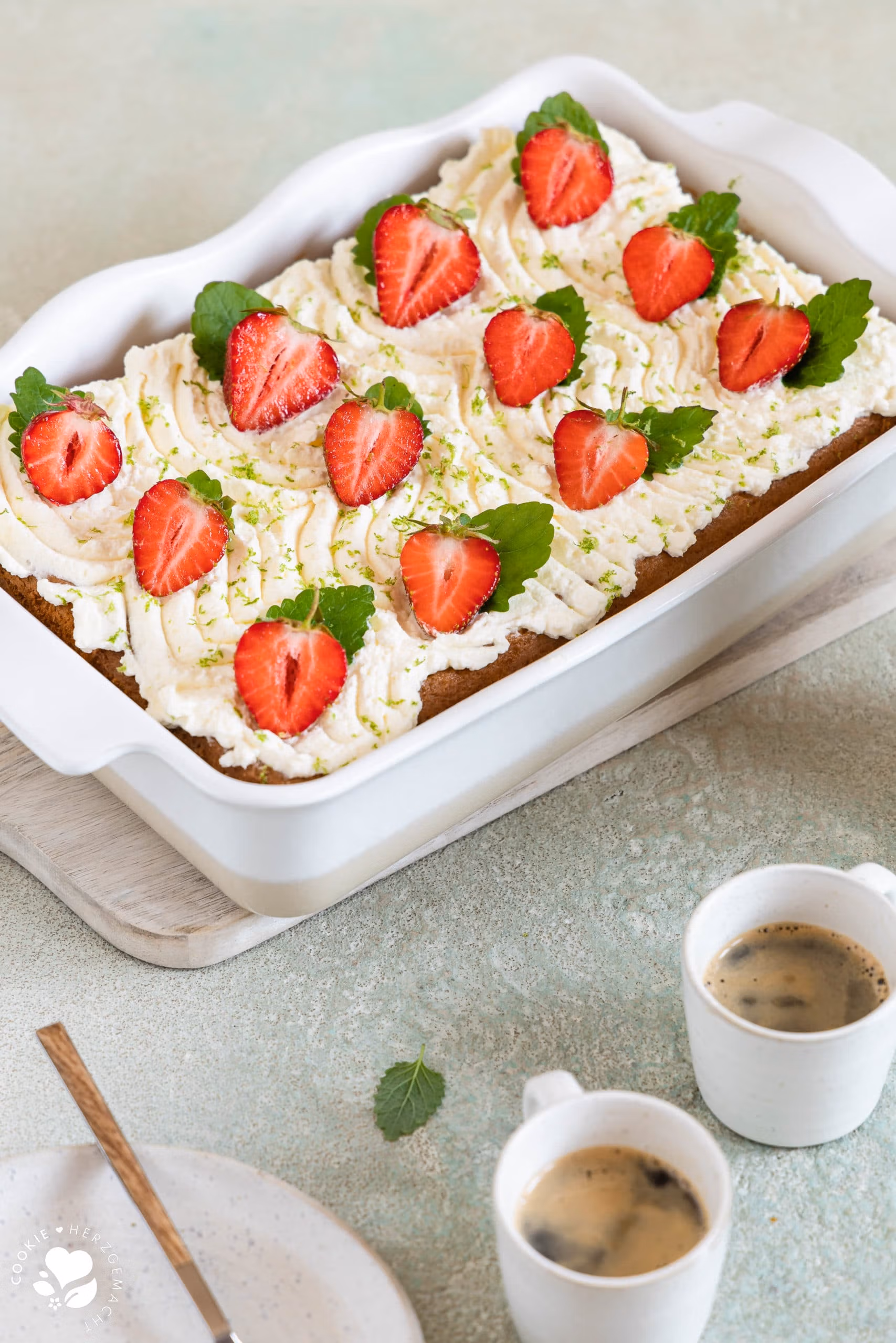 Ein Stück mexikanischer Tres leche Kuchen (Milchkuchen) mit Erdbeeren auf einem Teller mit Espressotassen und einer Backform mit Kuchen im Hintergrund.