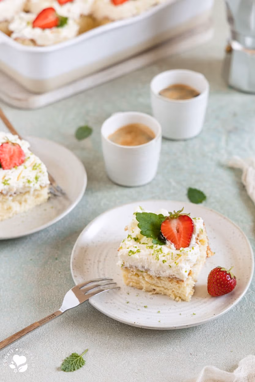 Ein Stück mexikanischer Tres leche Kuchen (Milchkuchen) mit Erdbeeren und Melisse auf einem Teller, einer Gabel im Vordergrund und einer Backform mit Kuchen und Espressotassen im Hintergrund.