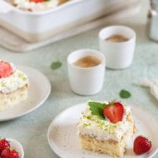 Ein Stück mexikanischer Tres leche Kuchen (Milchkuchen) mit Erdbeeren auf einem Teller mit Espressotassen und einer Backform mit Kuchen im Hintergrund.