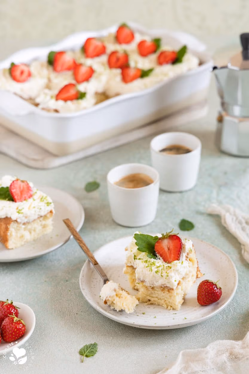 Ein Stück mexikanischer Tres leche Kuchen (Milchkuchen) mit Erdbeeren auf einem Teller mit Espressotassen und einer Backform mit Kuchen im Hintergrund.