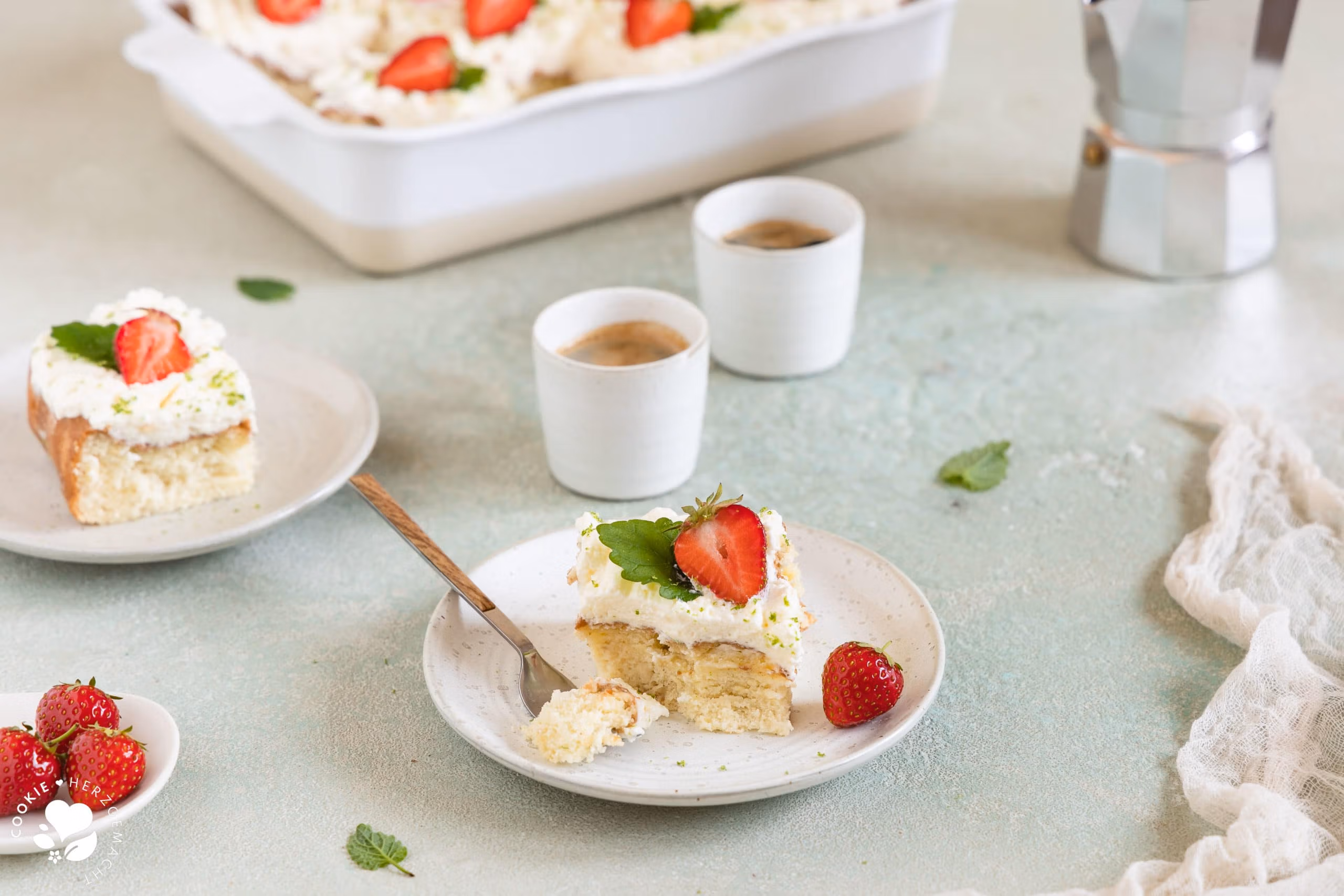 Ein Stück mexikanischer Tres leche Kuchen (Milchkuchen) mit Erdbeeren auf einem Teller mit Espressotassen und einer Backform mit Kuchen im Hintergrund.