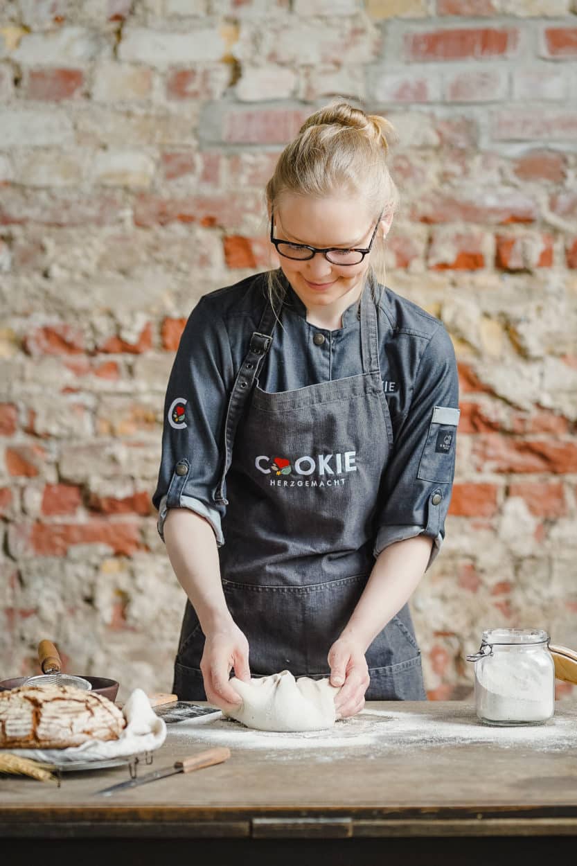 Sonja Bauer von Cookie und Co beim Formen von Brotteig auf einem Holztisch mit einer Backsteinmauer im Hintergrund.