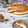 Ein Stück Karamell-Käsekuchen ohne Backen auf einem Teller mit einer Kuchengabel und dem Kuchen und Kaffeetassen im Hintergrund.