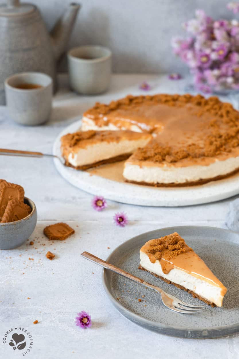 Ein Stück Karamell-Käsekuchen ohne Backen auf einem Teller mit einer Kuchengabel und dem Kuchen und Kaffeetassen und Blüten im Hintergrund.