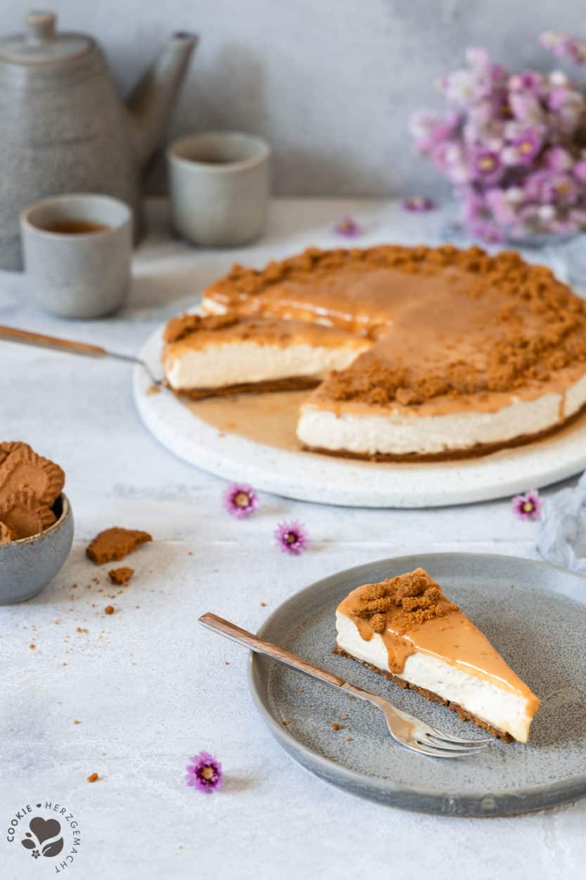 Ein Stück Karamell-Käsekuchen ohne Backen auf einem Teller mit einer Kuchengabel und dem Kuchen und Kaffeetassen im Hintergrund.