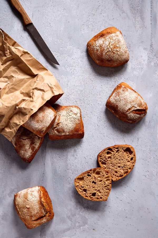Frisch gebackene, rustikale Vollkornbrötchen aufgebrochen auf einer hellen Betonplatte, daneben ein Brotmesser und eine Papiertüte.