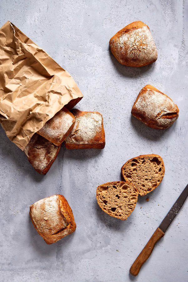 Aufgeschnittene Vollkornbrötchen mit lockerer Krume, daneben ein Brotmesser und weitere Brötchen auf einer strukturierten Oberfläche.