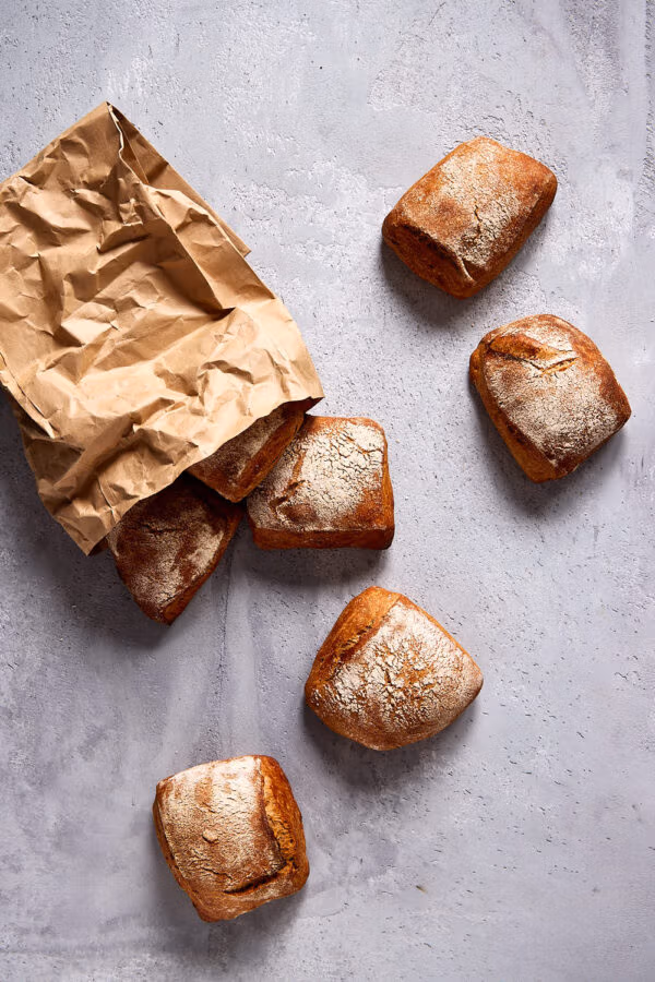 Mehrere goldbraune Vollkornbrötchen mit rustikaler Kruste, verteilt auf einer hellen Steinoberfläche mit Papiertüte im Bildausschnitt.