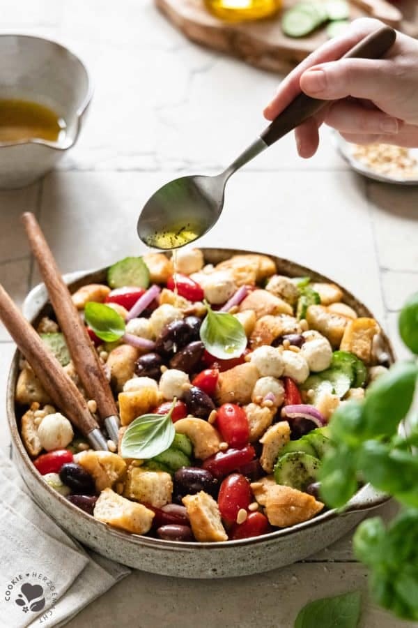 Italienischer Brotsalat „Panzanella“ in einer Salatschüssel, auf den das Dressing mit einem Löffel gegossen wird.