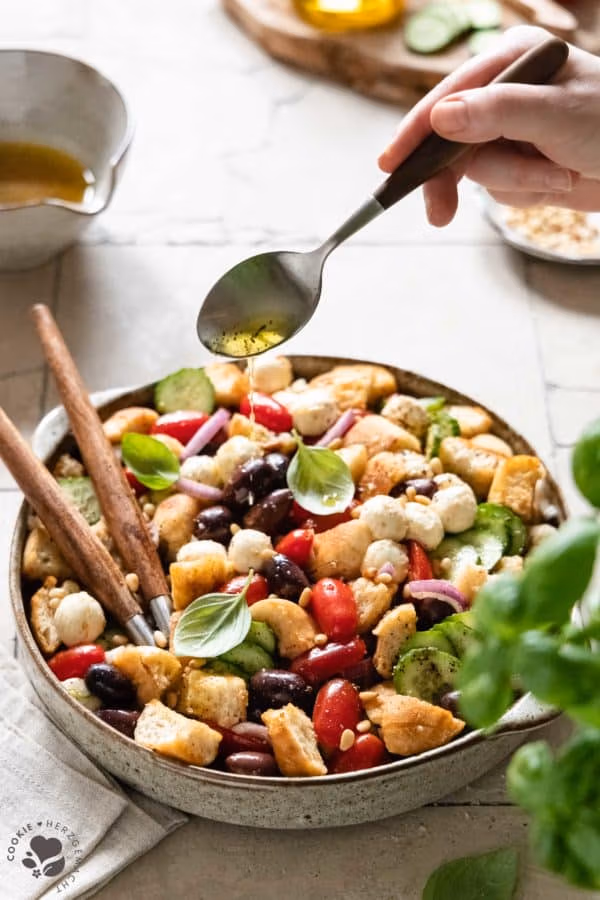 Italienischer Brotsalat „Panzanella“ in einer Salatschüssel, auf den das Dressing mit einem Löffel gegossen wird.