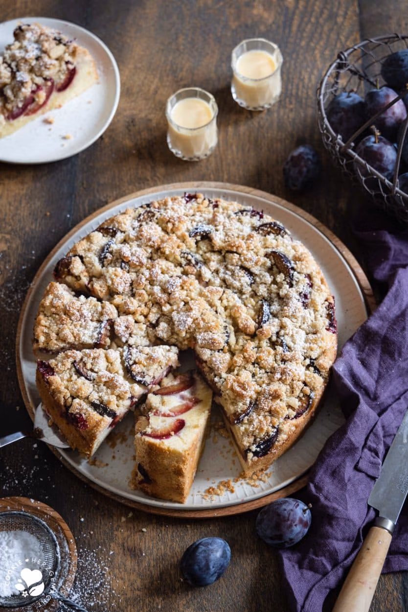 Ein Zwetschgenkuchen mit Streuseln auf einer Tortenplatte mit zwei Gläsern Eierlikör im Hintergrund und einem Korb mit Pflaumen.