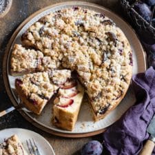 Ein Zwetschgenkuchen mit Streuseln auf einer Tortenplatte mit einem Puderzuckersieb im Hintergrund und einem Korb mit Pflaumen.