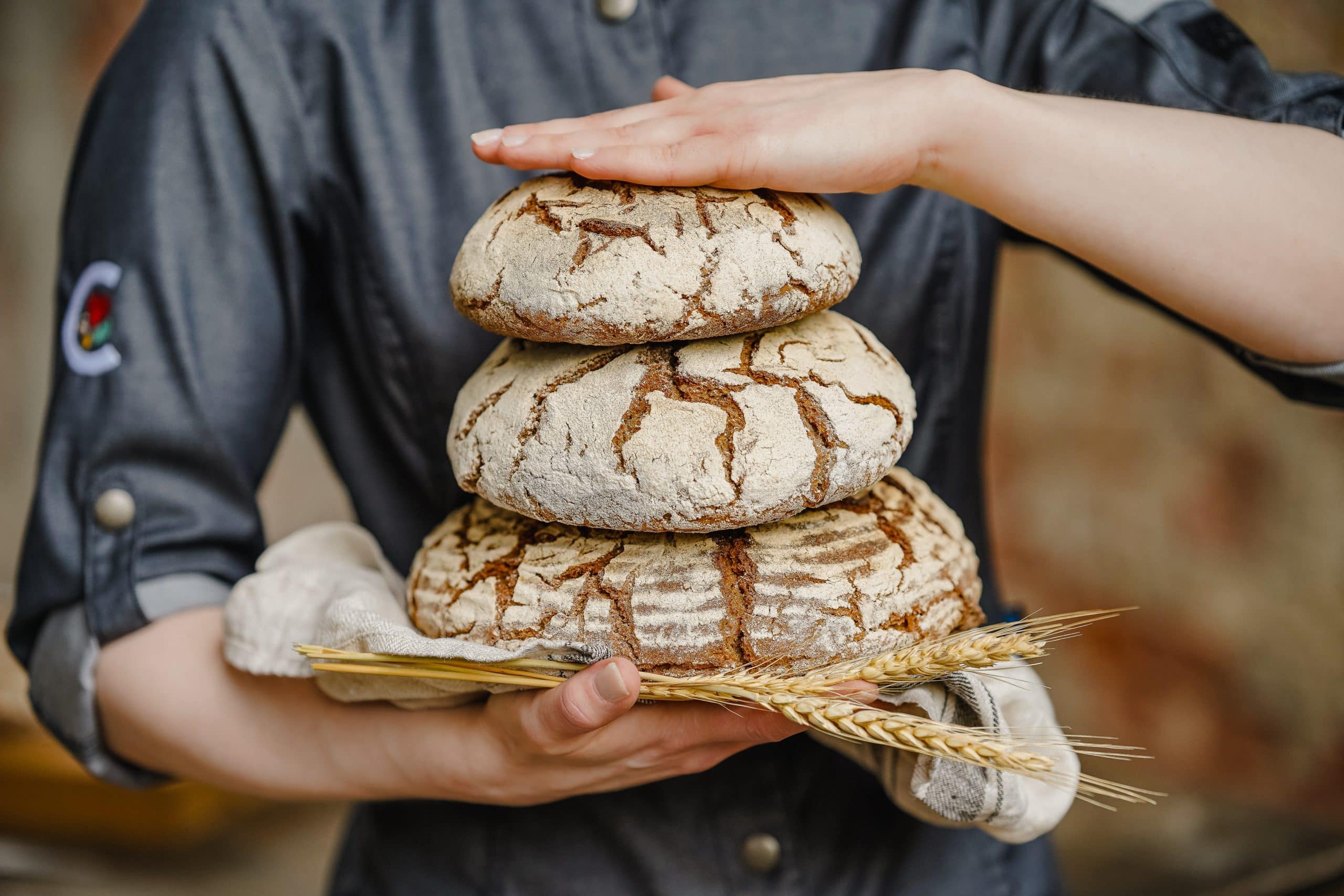 Drei Brotlaibe zwischen zwei Hände gestapelt, mit einem Leinentuch und Getreideähren darunter.