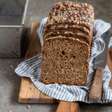 Ein Kastenbrot in Scheiben geschnitten auf einem Holzbrett mit einem blau gestreiften Tuch darunter und einer Backform im Hintergrund.