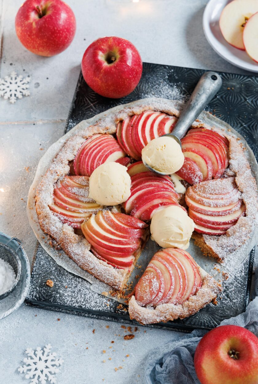 Eine Apfelgalette auf einem Backblech mit Vanilleeis und Äpfeln im Hintergrund.
