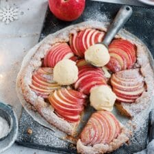 Eine Apfelgalette auf einem Backblech mit Vanilleeis und Äpfeln im Hintergrund.