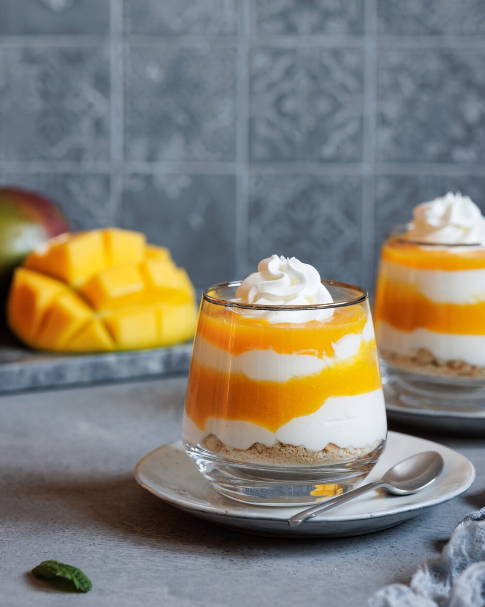 Schichtdessert mit Mango und Joghurt in einem Glas mit einer Sahnehaube und einem zweiten Glas im Hintergrund sowie einer aufgeschnittenen Mango.