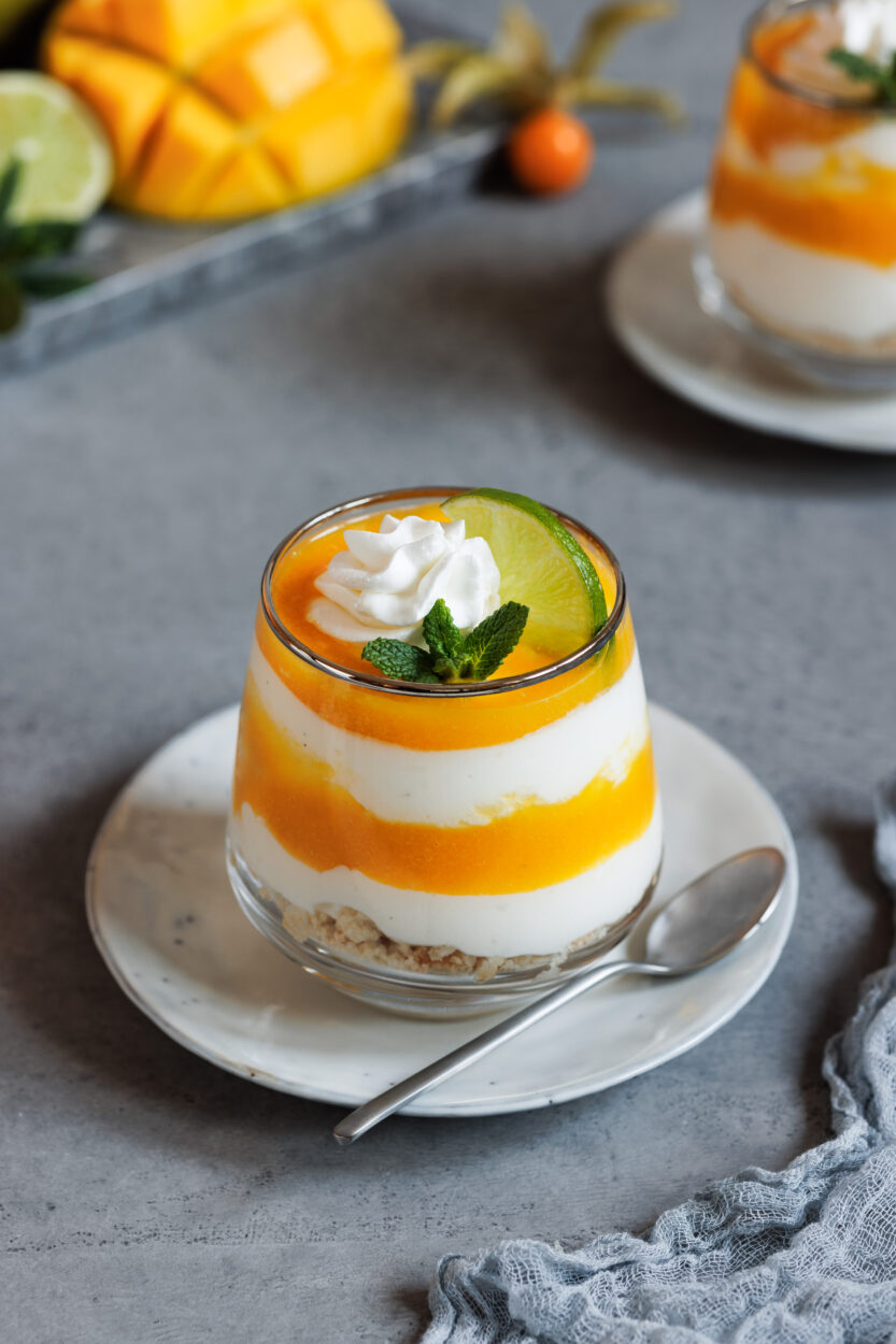 Mango-Dessert mit Joghurt in einem Glas, das auf einem Teller zusammen  mit einem Löffel steht. Im Hintergrund steht ein zweites Glas sowie eine aufgeschnittene Mango, Limette und Physalis.