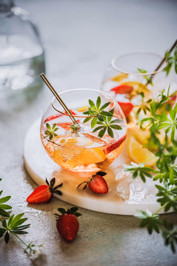 Maibowle mit Erdbeeren, Zitrone und Waldmeister in zwei Bowlegläsern auf einem Marmortablett mit Waldmeister und Erdbeeren im Vordergrund.