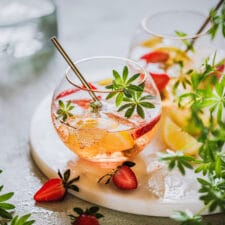 Maibowle mit Erdbeeren, Zitrone und Waldmeister in zwei Bowlegläsern auf einem Marmortablett mit Waldmeister und Erdbeeren im Vordergrund.