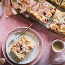 Ein Stück Rhabarber-Erdbeer-Kuchen mit Streuseln auf einem Teller mit zwei Tassen Kaffee daneben und einem Brett mit weiteren Kuchenstücken im Hintergrund.