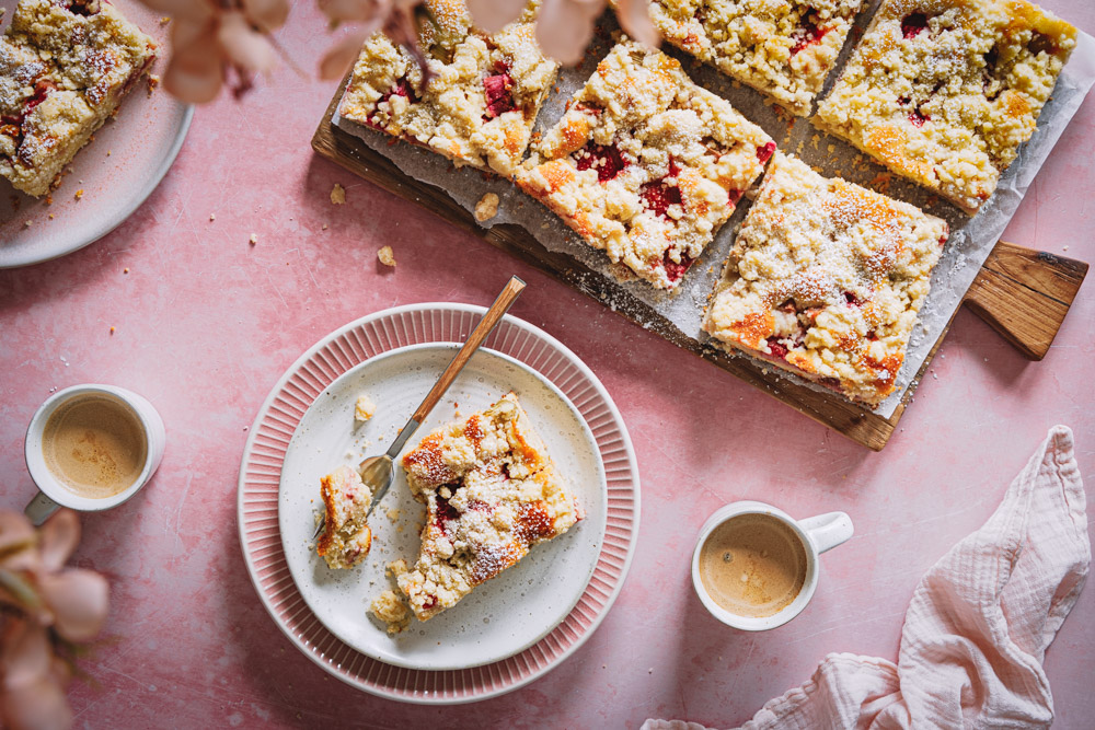 Ein Stück Erdbeer-Rhabarber-Kuchen mit Streuseln auf einem Teller mit zwei Tassen Kaffee daneben und einem Brett mit weiteren Kuchenstücken im Hintergrund. 