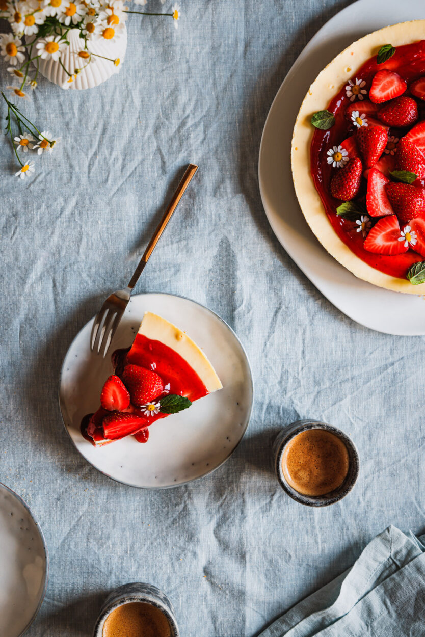 Ein Stück Erdbeer-Cheesecake auf einem Teller mit zwei Tassen Kaffee daneben und dem ganzen Käsekuchen und Blüten im Hintergrund. 