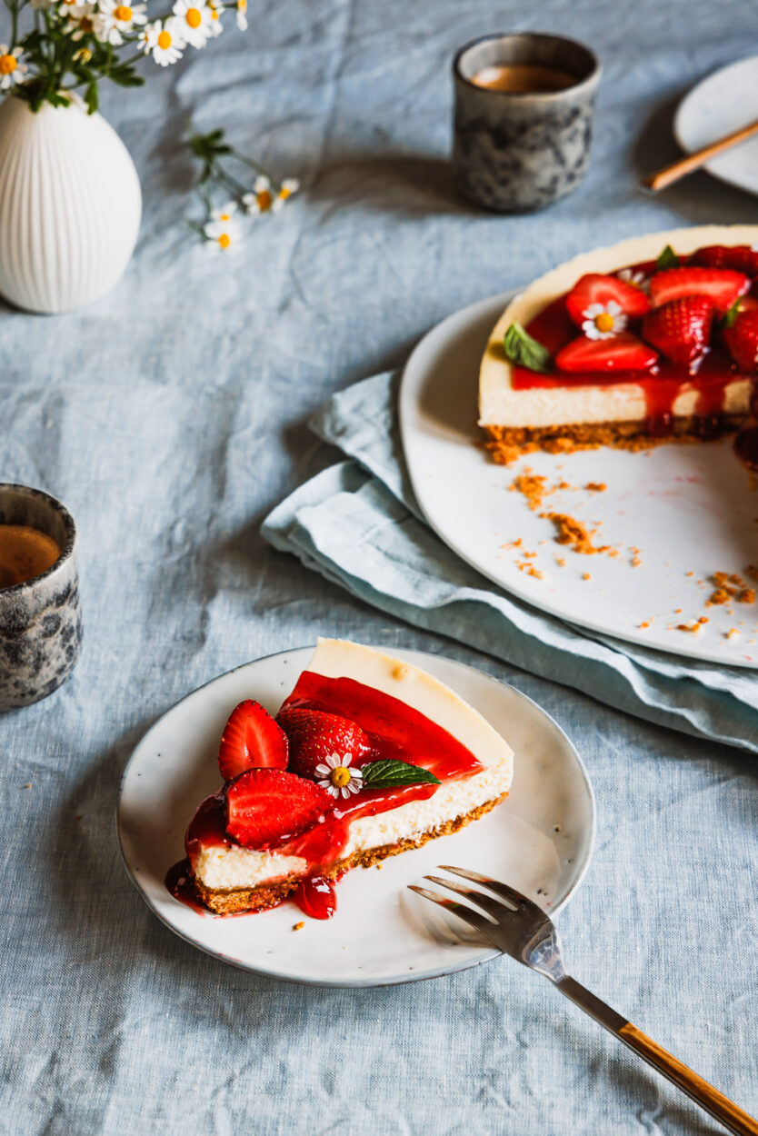 Ein Stück Erdbeer-Cheesecake auf einem Teller mit zwei Tassen Kaffee daneben und dem ganzen Käsekuchen und Blüten im Hintergrund.