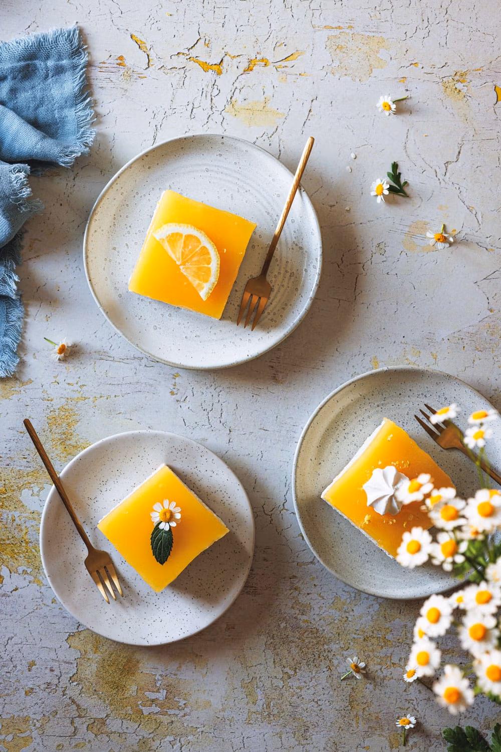 Lemon Cheesecake auf drei Tellern mit einer blauen Serviette und Blüten daneben.