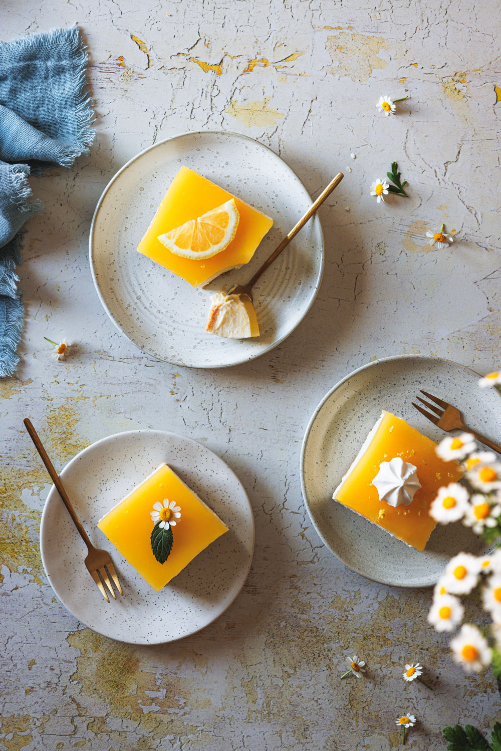 Lemon Cheesecake auf drei Tellern mit einer blauen Serviette und Blüten daneben.
