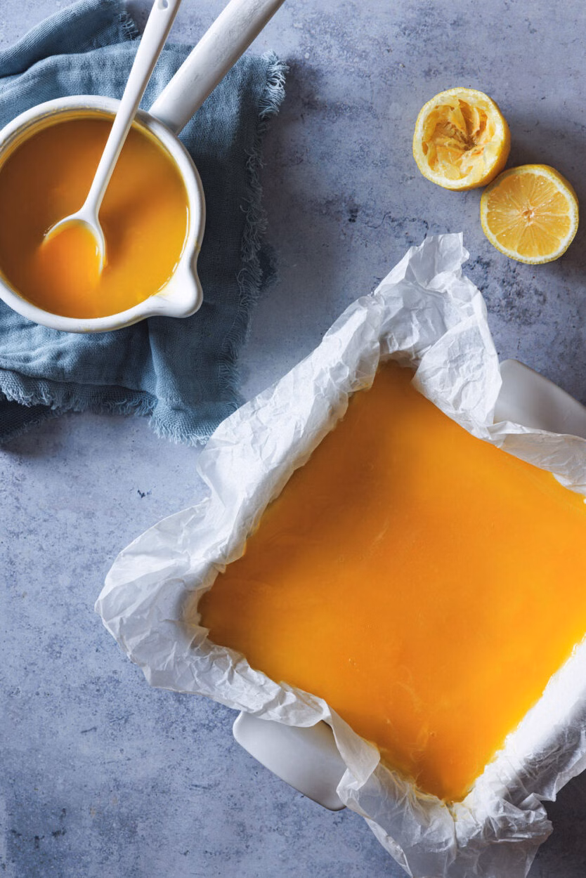 Zitronenkäsekuchen in einer Kuchenform mit Zitronen und einem Zopf mit dem Zitronentopping daneben. 