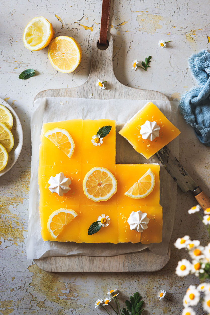 Lemon Cheesecake auf einem Holzbrett in Stücke geschnitten mit Zitronen und Blüten daneben.