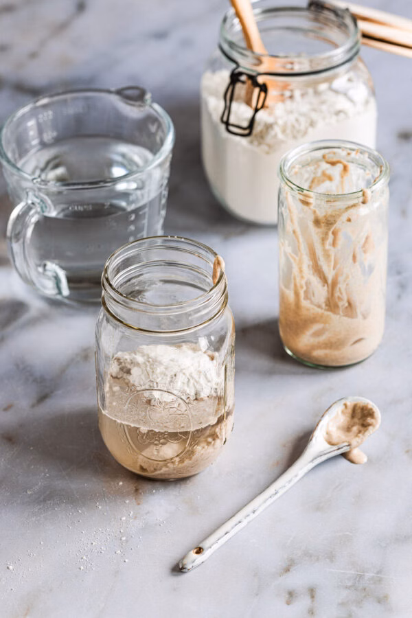 Sauerteig beim Auffrischen in einem Glas mit einem leeren Glas Anstellgut, einem Messbecher mit Wasser und einem Glas mit Mehl im Hintergrund.