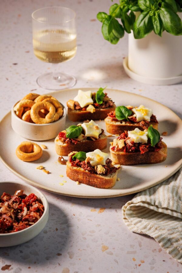 Bruschetta mit getrockneten Tomaten auf knusprigen Ciabatta-Scheiben, garniert mit Mozzarella-Sternen, frischen Basilikumblättern und zerbröselten Taralli auf einem Teller, dazu ein Glas Weißwein und ein Topf mit Basilikum im Hintergrund.