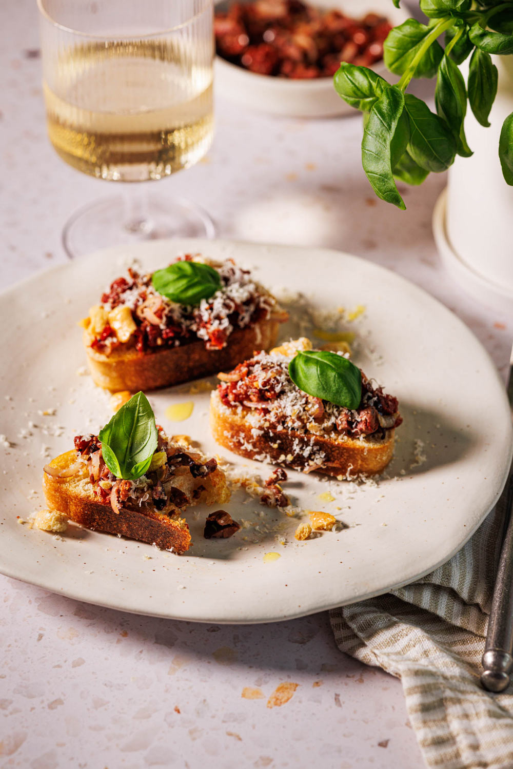 Bruschetta mit getrockneten Tomaten, Oliven und Schalotten auf gerösteten Ciabatta-Scheiben, garniert mit frischem Basilikum und geriebenem Parmesan auf einem Teller, im Hintergrund ein Glas Weißwein, eine Schale mit getrockneten Tomaten und ein Topf Basilikum.