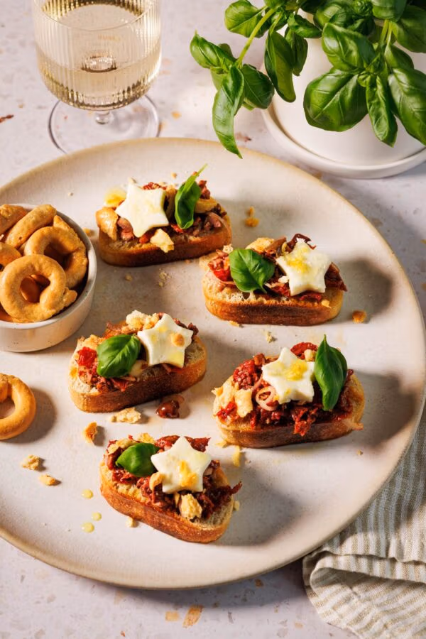 Bruschetta mit getrockneten Tomaten auf knusprigen Ciabatta-Scheiben, garniert mit Mozzarella-Sternen, frischen Basilikumblättern und zerbröselten Taralli auf einem Teller, dazu ein Glas Weißwein und ein Topf mit Basilikum im Hintergrund.