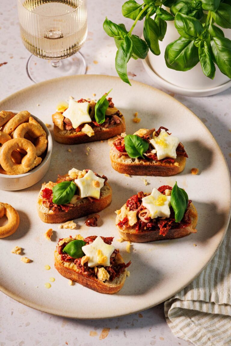 Bruschetta mit getrockneten Tomaten auf knusprigen Ciabatta-Scheiben, garniert mit Mozzarella-Sternen, frischen Basilikumblättern und zerbröselten Taralli auf einem Teller, dazu ein Glas Weißwein und ein Topf mit Basilikum im Hintergrund.