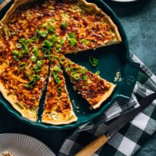 Eine frisch gebackene Quiche Lorraine in einer grünen Tarteform, aufgeschnitten und garniert mit geschnittenen Frühlingszwiebeln. Einige Stücke sind entnommen und auf weißen Tellern serviert. Daneben liegt ein Messer auf einer grün-weiß karierten Serviette.