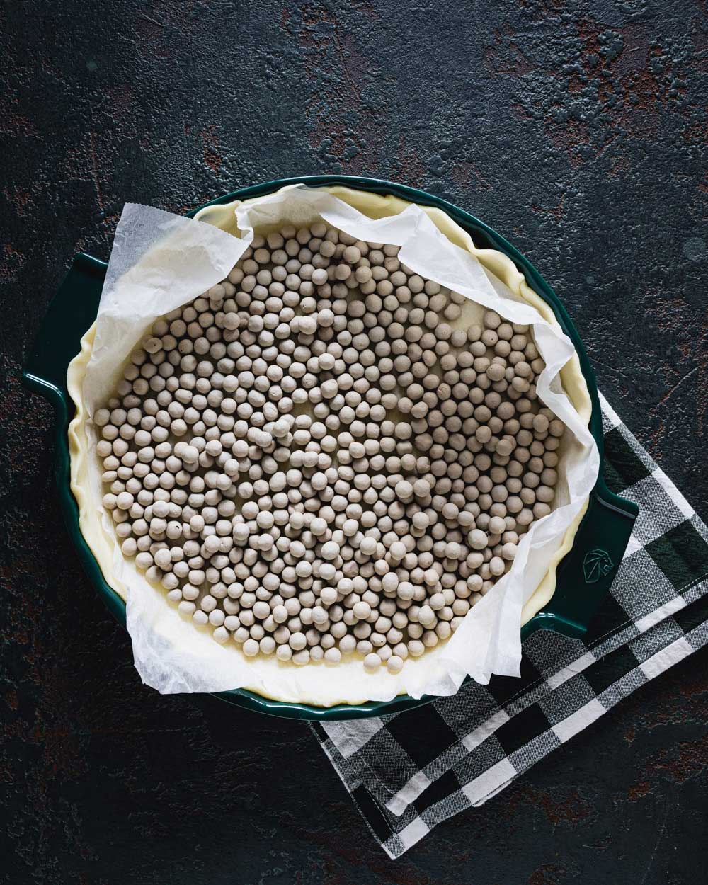 Quiche-Teig in einer grünen Tarteform, mit Backpapier ausgelegt und mit Blindback-Kugeln beschwert, bereit zum Vorbacken auf einem dunklen Hintergrund mit einem schwarz-weiß karierten Geschirrtuch darunter.