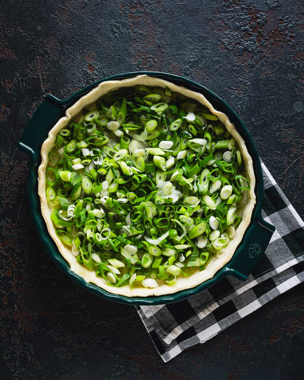 Eine grüne Tarteform mit vorgebackenem Quiche-Teig, der mit frisch geschnittenen Lauchzwiebeln gefüllt ist, auf einem dunklen Hintergrund mit einem schwarz-weiß karierten Geschirrtuch darunter.