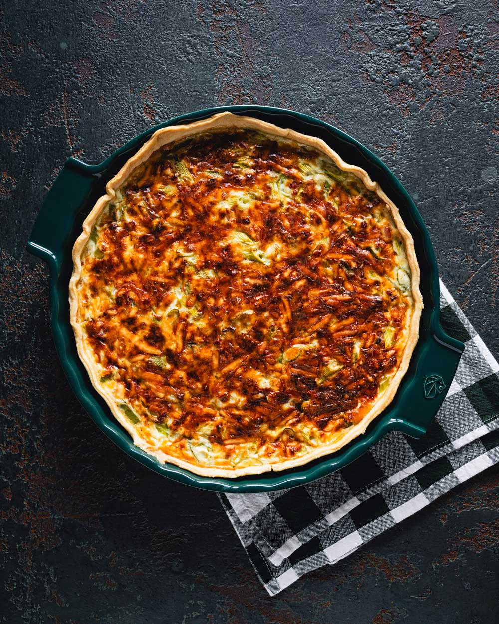 Fertig gebackene Quiche Lorraine in einer grünen Tarteform, mit goldbrauner, knuspriger Käsekruste, auf einem dunklen Hintergrund mit einem schwarz-weiß karierten Geschirrtuch darunter.