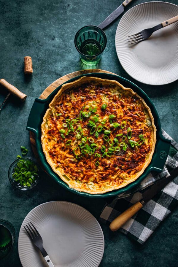 Frisch gebackene Quiche Lorraine in einer grünen Tarteform, garniert mit Frühlingszwiebeln, auf einem Tisch mit gedeckten Tellern, Gabeln und einem grünen Glas. Im Hintergrund liegen ein Korkenzieher und ein Korken.