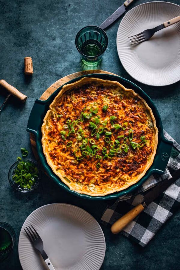 Frisch gebackene Quiche Lorraine in einer grünen Tarteform, garniert mit Frühlingszwiebeln, auf einem Tisch mit gedeckten Tellern, Gabeln und einem grünen Glas. Im Hintergrund liegen ein Korkenzieher und ein Korken.