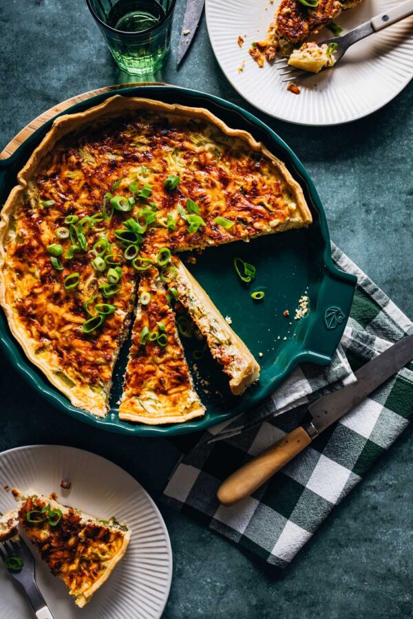 Frisch gebackene Quiche Lorraine in einer grünen Tarteform, aufgeschnitten und garniert mit Frühlingszwiebeln. Ein Stück liegt auf einem weißen Teller mit einer Gabel, daneben ein Messer auf einem schwarz-weiß karierten Geschirrtuch.